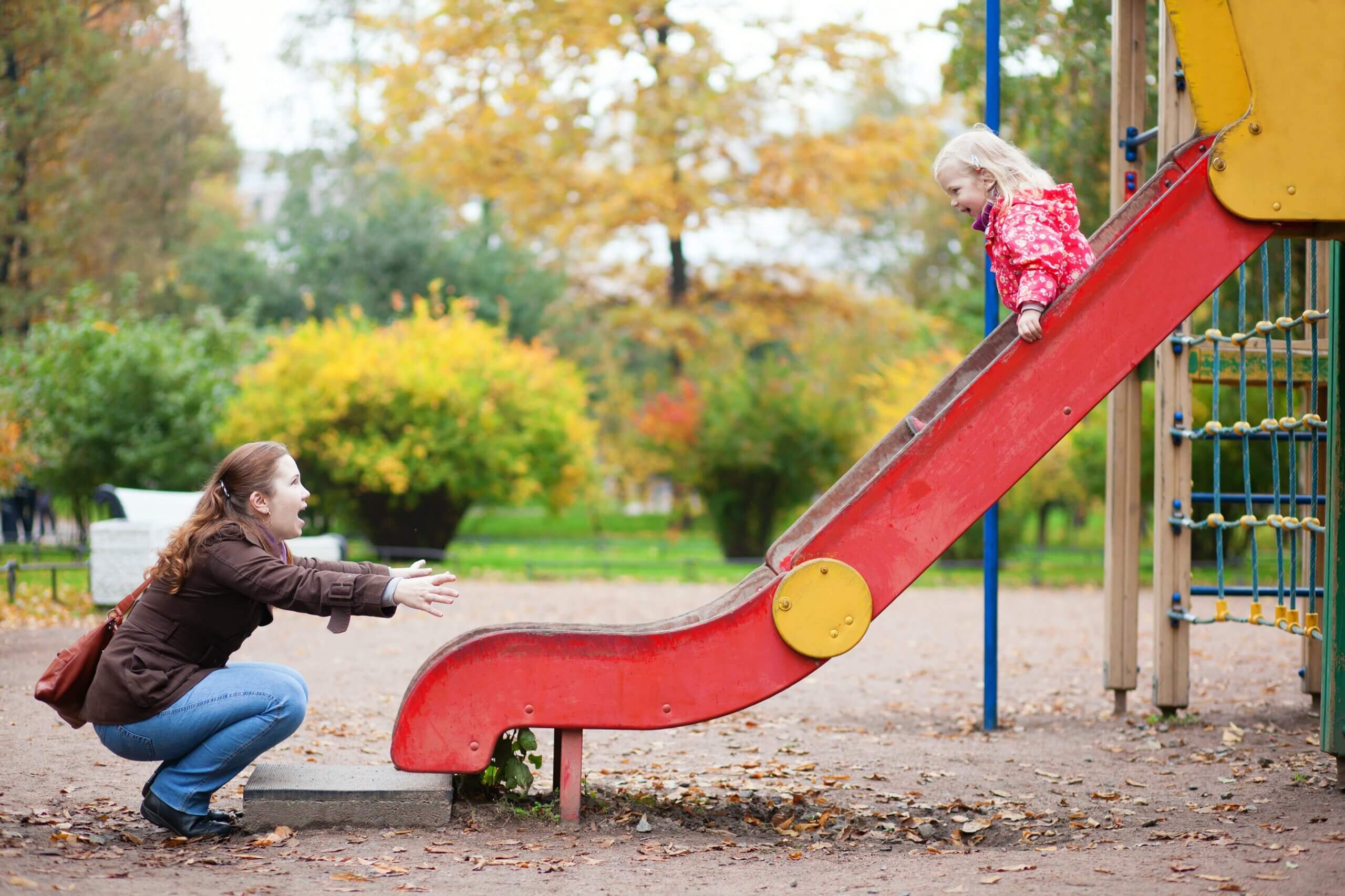 A girl on a slide and a mom waiting for her at the bottom with open arms